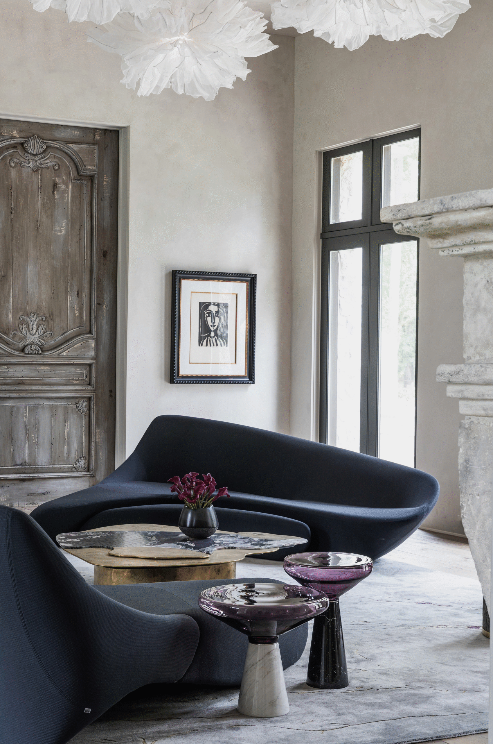 Memorial residence by Nina Magon Studio featuring a sculptural midnight-blue curved sofa, artisanal stone side tables, a marble fireplace, and historic wooden doors layered with gallery artwork.